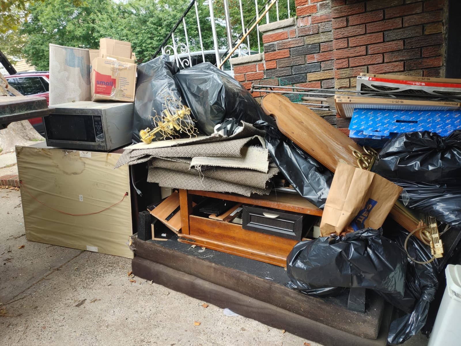 Curbside pile with a microwave, rolled carpet, dresser, wooden furniture and bagged trash.